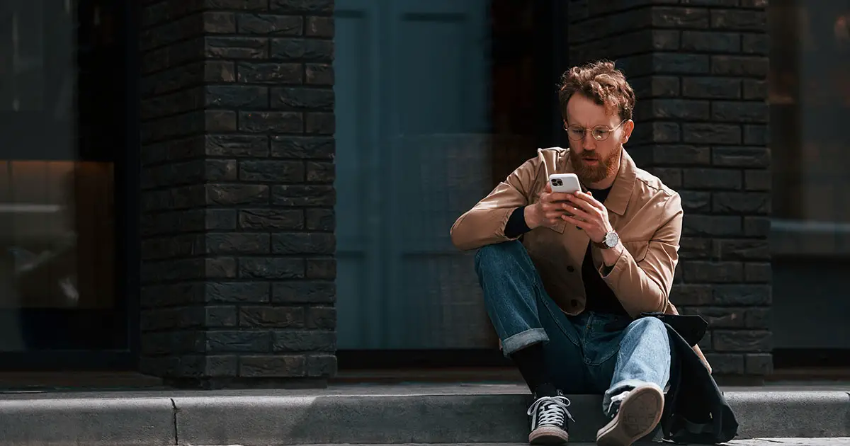 Stylish man wearing jeans looks at smartphone while sitting on sidewalk.