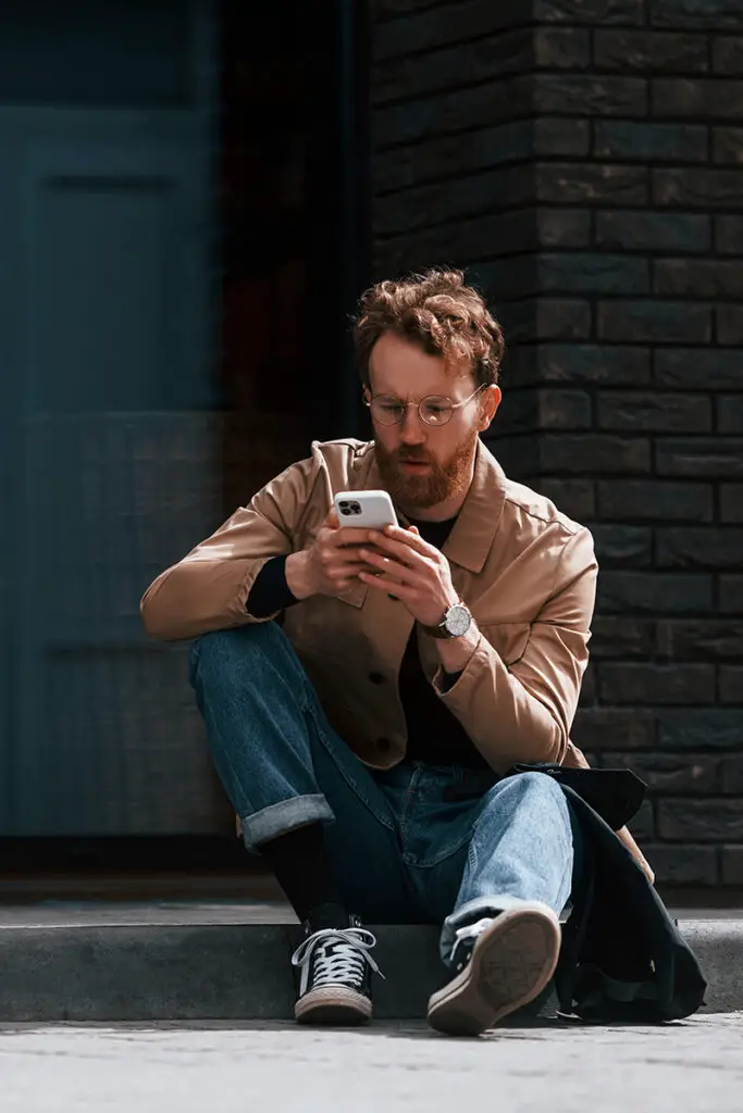 Stylish man wearing jeans sits on sidewalk looking at smartphone.