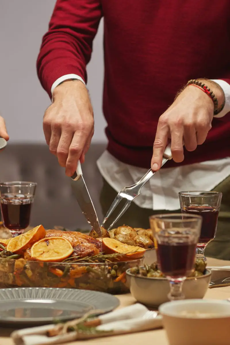 Man wearing stylish Thanksgiving outfit cuts turkey at table.